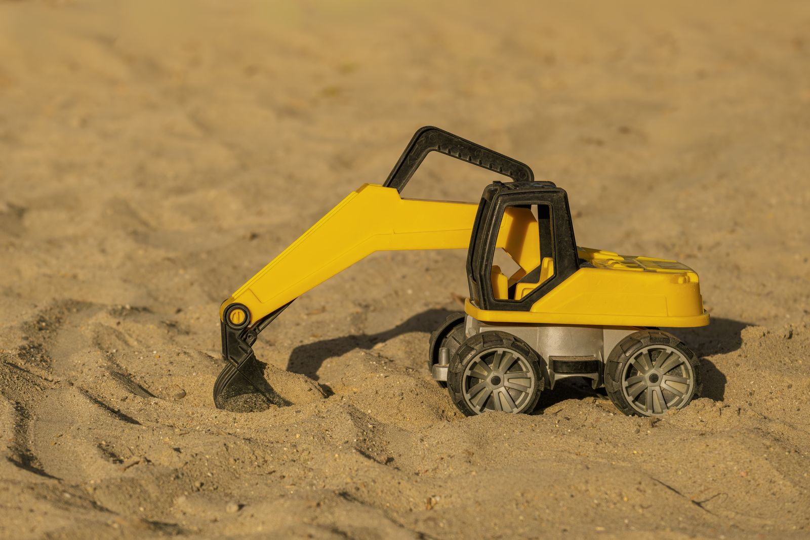 Close-up of a yellow toy excavator on a sandy surface under bright sunlight.